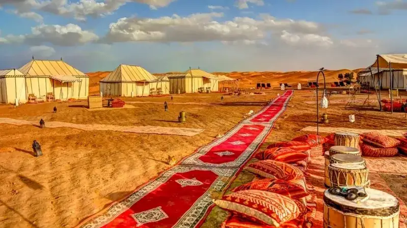 A luxury camp in the Moroccan desert featuring tents set on a vibrant carpet against a sandy landscape.